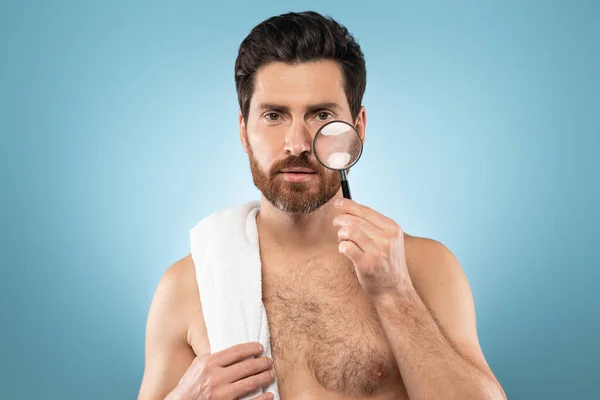 Handsome man using magnifying glass, looking for wrinkles on face on blue studio background. Skin care, dermatology, cosmetics and healthy hair and body