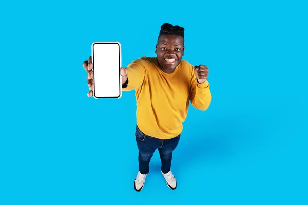 兴奋的黑人男子用白屏演示空白智能手机庆祝成功 快乐的非洲裔美国人享受新的移动应用程序或在线服务 站在蓝色背景上 — 图库照片