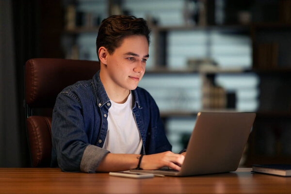 Handsome positive young guy startuper sitting at table, working at dark office, using laptop, typing on pc keyboard, sending emails, coding, testing newest computer software, copy space