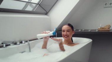 Vücut bakımı ve hijyen. Genç mutlu bayan sıcak köpüklü küvette dinleniyor, eline şampuan döküyor ve saçlarını yıkıyor, takip çekimi, yavaş çekim, boş alan.