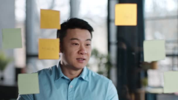 成功した仕事の計画 オフィスのガラスの壁にメモを貼り付ける積極的な中年アジアのマネージャーは 新しいプロジェクト戦略を準備し ショットを追跡し スローモーション — ストック動画