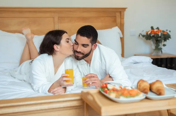 新婚旅行ロマンス 頬に夫とキスを愛する妻はジュースを飲んで モダンなホテルスイート屋内に横たわっているベッドでロマンチックな朝食をお楽しみください バスローブを着て 選択的フォーカス — ストック写真