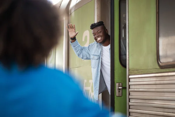 Cheerful Black Man Waving Hand To Girlfriend While Standing In Train Door At Railway Station, Joyful Handsome African American Guy Greeting His Spouse After Arrival Or Saying Bye Before Leaving
