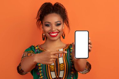 Neşeli gülümseyen, parlak makyaj yapan güzel siyah kadın ve geleneksel Afrika kostümü giyen, beyaz ekranlı modern cep telefonunu gösteren, online teklif için model, turuncu arka plan