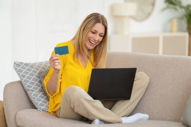 Gülen bin yıllık Kafkasyalı sarışın alışveriş meraklısı kadın, dizüstü bilgisayarında kanepenin üzerinde kredi kartı gösteriyor, online alışveriş yapıyor ve oturma odasının içinde sipariş veriyor. Büyük indirim, nakit para ve evde bankacılık.