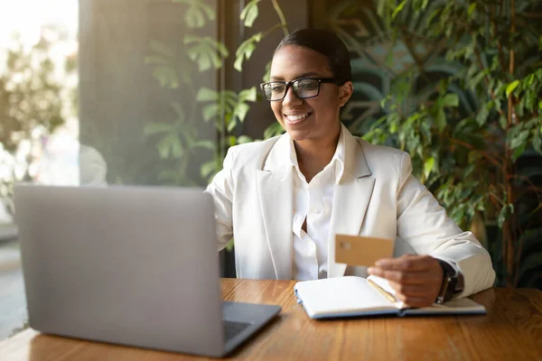 Happy millennial black businesswoman in white suit and glasses uses laptop and credit card in cafe office with green plants interior. Gadget for business, online shopping, finance and money