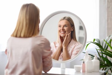 Olgun Güzellik. Orta yaşlı çekici bir kadının portresi aynadaki yansımasına gülümsüyor, güzel bir kadın evde Skincare rutini yaparken görünüşünün keyfini çıkarıyor, seçici bir şekilde odaklanıyor.