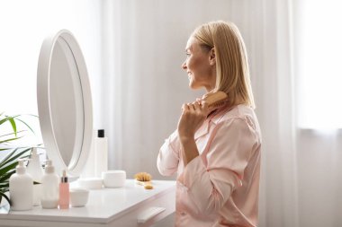 Güzel, orta yaşlı bir kadın, yatak odasında giyinme masasında otururken sarı saçlarını tarayıp tarıyor, mutlu, olgun bir kadın, yansımasına ve gülüşüne bakıyor, yandan bakıyor.