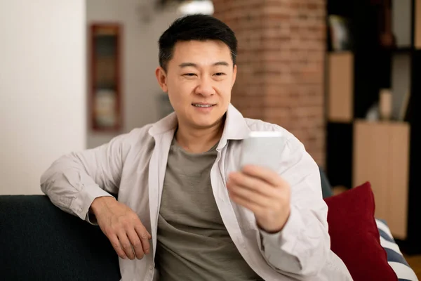 在家中沙发上休息 在手机上与韩国男性聊天或上网购物时 用手机微笑的亚洲中年男人 — 图库照片