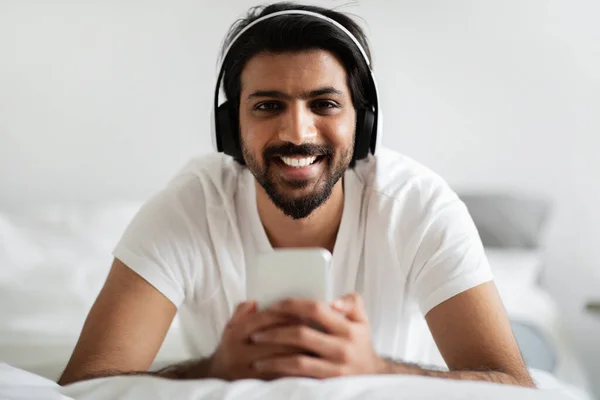 Wesoły Młody Człowiek Bliskiego Wschodu Białej Koszulce Słuchawkach Smartfonie Leży — Zdjęcie stockowe