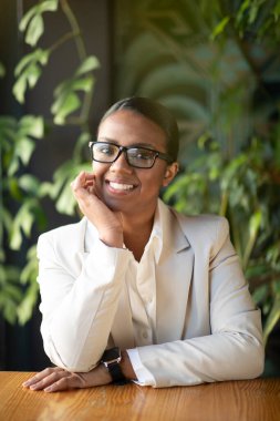 Beyaz takım elbiseli Afro-Amerikan iş kadını gülümsüyor ve kafenin içinde yeşil bitkilerle gözlük takıyor, dikey, dikey, yakın. Modern iş, iş ve yaşam tarzı