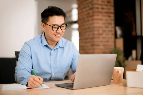 Szczęśliwy Azjatycki Biznesmen Robi Notatki Siedzi Stół Laptopem Oglądać Webinarium — Zdjęcie stockowe