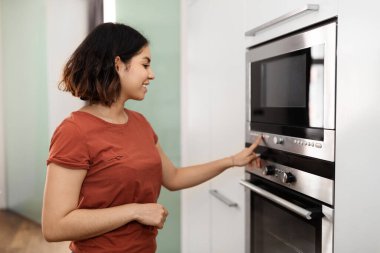 Güzel bir Arap kadının mutfakta elektrikli fırını açması Orta Doğulu bir bayanın parmaklarıyla kontrol paneline dokunması, evde yemek hazırlarken modern mikrodalgada pişirme rejimini seçmesi.