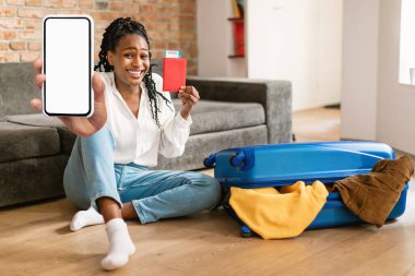 Rezervasyon uygulaması. Büyük boş akıllı telefon gösteren, elinde biletlerle pasaport tutan, bavulunun yanında oturan, tatile çıkmaya hazır mutlu siyah kadın.