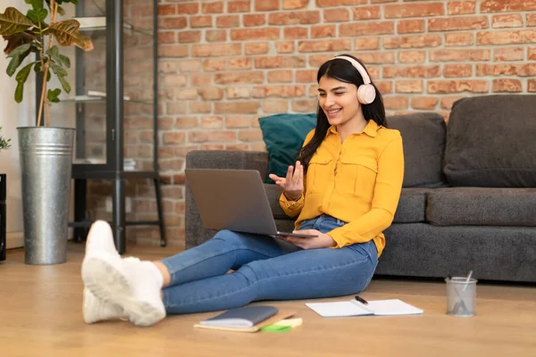 Happy young spanish lady in wireless headphones talking at laptop webcam, having video call in living room, studying online. Webinar and online lesson at home