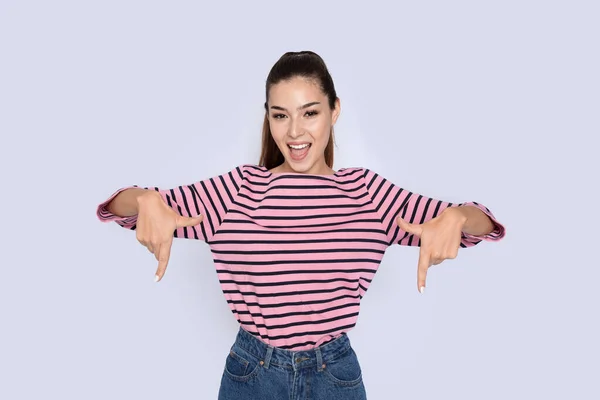 You have to see it. Emotional excited pretty brunette young lating woman wearing casual outfit pointing down with both hands, posing on grey studio background, copy space