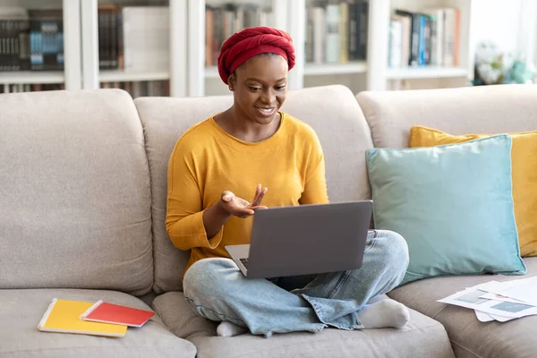 Pretty positive young black woman employee have video call with colleagues, working from home, sitting on couch, wearing casual outfit and african turban, using laptop, copy space. Remote job