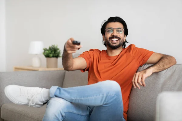 戴眼镜快乐的印度年轻男性坐在沙发上 带着遥控器 看电视 在客厅里享受闲暇时光 家庭娱乐 — 图库照片