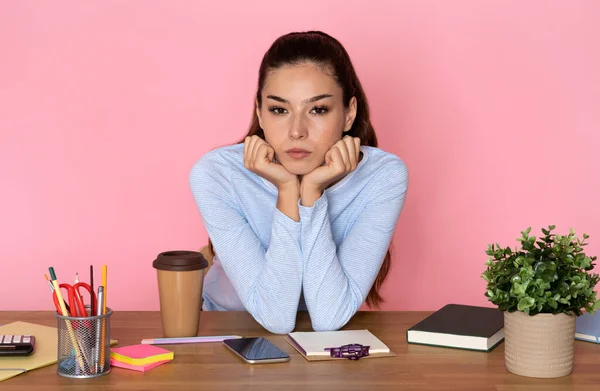 stock image Unloved job. Unhappy bored exhausted pretty hispanic young woman in smart casual outfit employee sitting at table full of stationery, cell phone, looking at camera, pink studio background