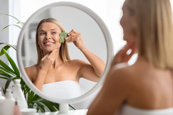 漂亮的中年妇女坐在镜子前 用瓜沙工具做面部按摩 迷人的成熟女士洗澡后做美容治疗 有选择地注重反思 — 图库照片