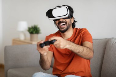 VR gözlükleri takmış neşeli, orta doğulu bir genç. Oturma odasının içinde joystick ile bilgisayarda gündelik oyunlar oynuyor. Boş zamanlardaki oyuncu, evdeki modern teknoloji.