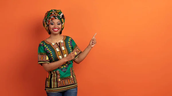 いい取引だ 広告や笑顔のための無料スペースを指している伝統的な明るいアフリカの衣装で陽気魅力的な若い黒人女性 オレンジのスタジオの背景に孤立 ウェブバナー — ストック写真