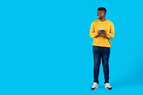 移动报价 笑着拿着智能手机的黑人年轻人放眼远方的复制空间 快乐的千年非裔美国人 用蓝色背景 全长的电话站着 — 图库照片