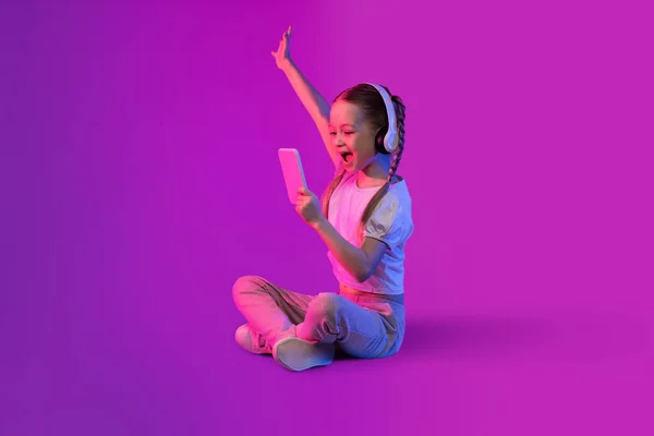 在霓虹灯的明亮背景下 用现代无线耳机和智能手机 看着手机屏幕和手势 查看最新的移动应用程序 兴奋而又美丽的年轻金发姑娘 — 图库照片