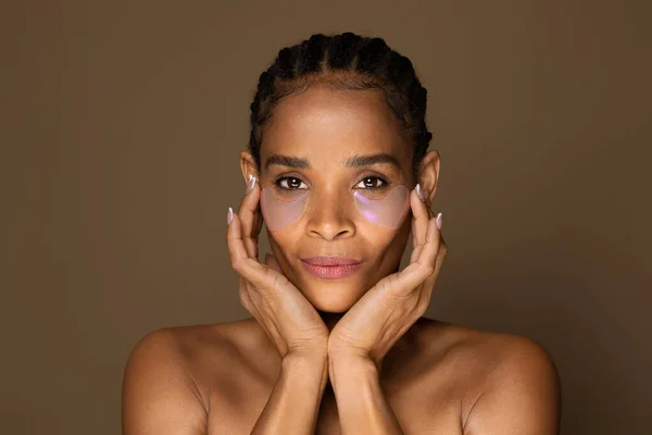 Beautiful black middle aged woman with eye patches touching face, enjoying procedures, posing isolated on brown background. Skin care, natural beauty, spa treatment concept