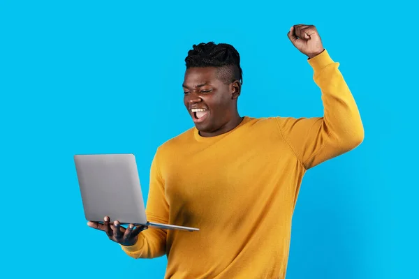 Joyful Black Man Celebrating Success With Laptop, Raising Clenched Fist And Exclaiming With Excitement, Happy African American Guy Holding Computer, Cheering Victory And Luck Over Blue Background