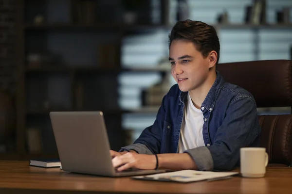 Happy inspired hard-working handsome young businessman in casual outfit sitting at workdesk late at night, typing on laptop keyboard, working in dark empty office, drinking coffee, copy space