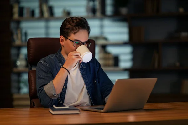 勤劳的年轻人在家里办公室工作到很晚 穿着休闲装和眼镜坐在黑暗房间的办公桌前 使用现代笔记本电脑 喝咖啡 复制空间 — 图库照片