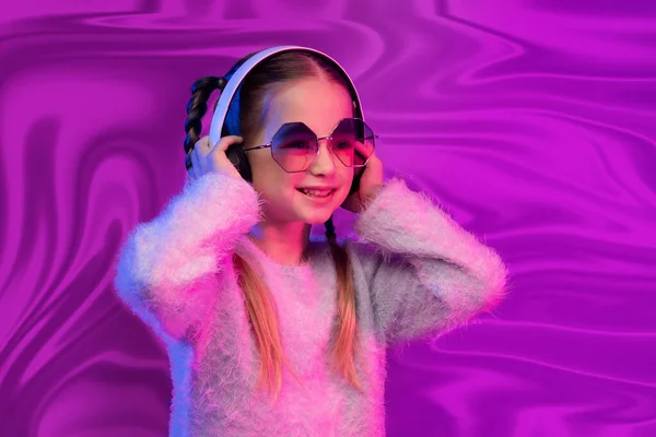 甘いです可愛いです可愛いですブロンドの女の子学校 サングラスで現代の無線ヘッドフォンを使用して 幸せな子供は音楽を聞いて 楽しみますピンク虹彩ホログラフィック箔の背景 — ストック写真