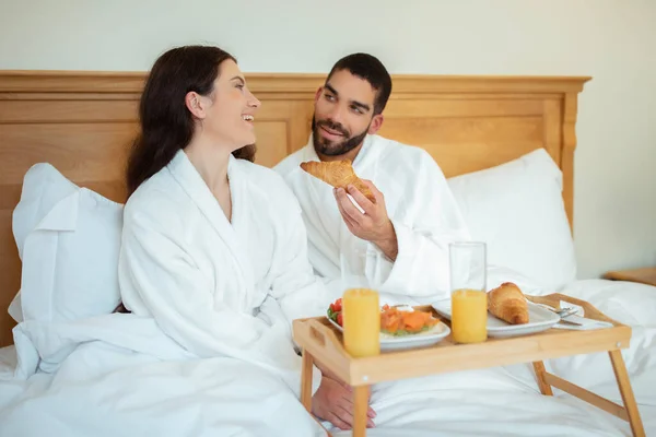朝はベッドの中でロマンチックな朝食をお楽しみください 夫はホテルスイートでテーブルトレイでポーズ妻にクロワッサンを提供しています 宿泊室サービスコンセプト — ストック写真
