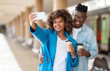 Neşeli siyah çift tren istasyonunda beklerken akıllı telefondan selfie çekiyor, Afro-Amerikalı genç bir adam ve kadın ellerinde kahve tutuyor ve kameraya gülümsüyor.