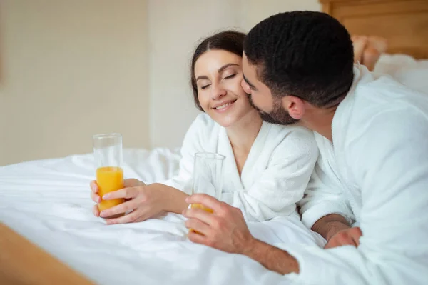 ロマンチックな休暇 男は現代のホテルの部屋の屋内で白いバスローブを着てオレンジジュースを保持ベッドに横たわっている女性のキス 新婚旅行を楽しむベッドでリラックスした多文化カップル — ストック写真