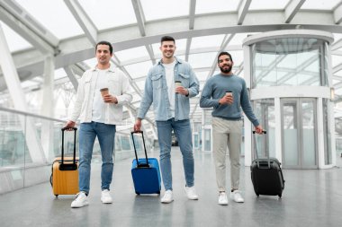 Elinde kağıt kahve bardaklarıyla gezen, havaalanı terminalinde seyahat çantalarıyla poz veren, kameraya gülümseyen üç turistin uzun pozu. Tatil ve Turizm Konsepti