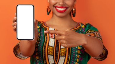 Afrikalı kostümlü siyahi kadın elinde beyaz ekranlı modern cep telefonu elinde tanınmayan bir kadın elindeki akıllı telefonu işaret ediyor reklam için model, turuncu stüdyo arka planı.
