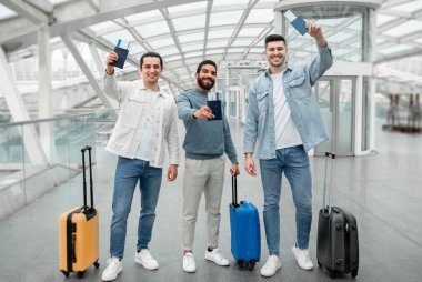 Ucuz Uçuş Teklifi. Pasaportlarını elinde biletlerle gezen ve modern havaalanında kameraya gülümseyen üç neşeli adam. Tam Uzunluk