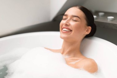 Gözlerini kapayan kadın köpükle banyo yapıyor suda yatıyor ve modern banyoda dinleniyor. Muhteşem Genç Bayan Banyo Yapan Spa Günü. Yurt İçi Rahatlama, Güzellik ve Sağlık