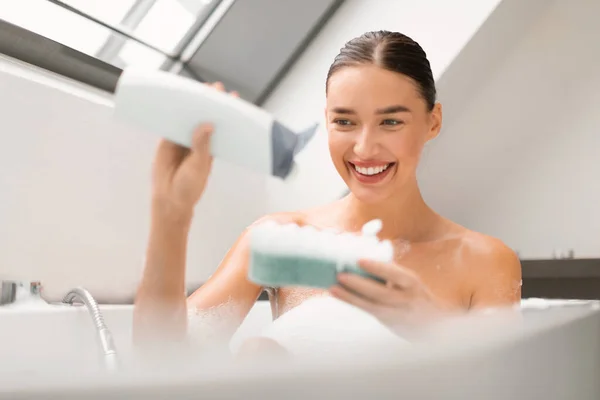 笑顔の女性は自宅でバスルームに座ってスポンジ洗濯ボディにバスタブとスクイーズシャワージェルを取ります 化粧品店内で衛生ルーチン入浴を楽しむ女性 選択的フォーカス — ストック写真