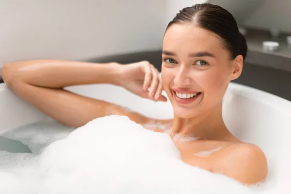 泡でお風呂に入る可愛い女性の美肖像画 自宅で浴室の浴槽で水にカメラをポーズを見て微笑む リラクゼーション 入浴化粧品コンセプト — ストック写真