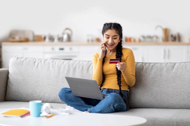 Çevrimiçi sipariş. Kredi kartı tutan ve cep telefonuyla konuşan mutlu Hintli kadın evde koltukta otururken, doğu kadını dizüstü bilgisayar kullanıyor ve müşteri destek müdürüyle sohbet ediyor.