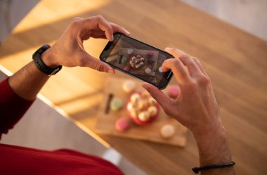 Hamur işlerinin fotoğrafını çeken tanınmamış bir adam ahşap masanın üzerinde çeşitli pastalar yapıyor cep telefonu kullanıyor, yemek blogcusu nüfuzunu kesiyor lezzetli tatlılar çekiyor, uzay boşluğunu kopyalıyor.