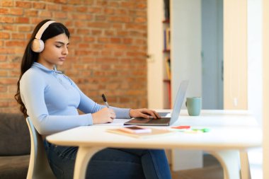 Uzaktan öğrenme konsepti. Genç bayan dizüstü bilgisayarla çalışıyor, notlar alıyor ve online ödev yapıyor, masada oturuyor, kablosuz kulaklık takıyor, boş yer arıyor.