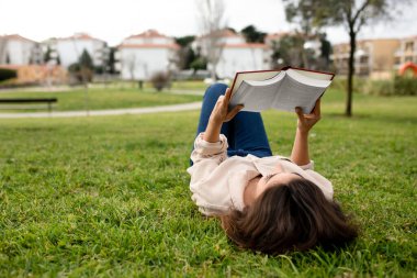 Milenyum Kafkasyalı esmer kız öğrenci çimlerin üzerinde uzanıyor, üniversite kampüsünde kitap okuyor, dinleniyor, dinleniyor ve kırılıyor, dışarıda. Üniversitede hobi, öğrenim, bilgi, eğitim ve yaşam tarzı