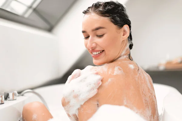 Happy Woman Washing Her Body Touching Smooth Skin With Applied Shower Gel Taking Bath Indoors. Lady Rubbing Shoulder Covered With Foam Bathing In Bathroom. Selective Focus