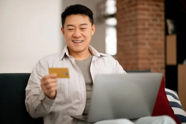 Happy korean man relaxing on couch in living room, using credit card and modern laptop, male customer shopping on Internet, banking online from home
