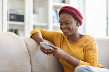 Mutlu, güzel, Afro-Amerikalı bir kadın günlük kıyafetler giyiyor ve geleneksel türban takıyor. Evdeki koltukta oturuyor, akıllı telefon kullanıyor, gülümsüyor, cep telefonu uygulamasında erkeklerle sohbet ediyor, uzay kopyaları kopyalıyor.