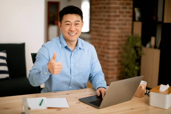 I like. Happy asian middle aged man sitting at workplace at home office, using laptop and showing thumb up, enjoying remote work, looking and smiling at camera